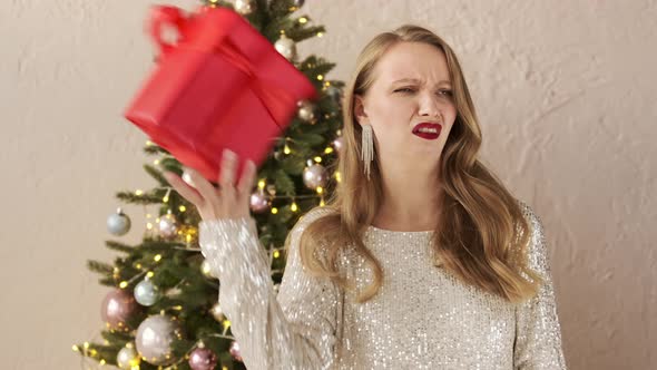 Festive woman in a shiny evening dress near Christmas tree gets a bad gift and throws it away alt