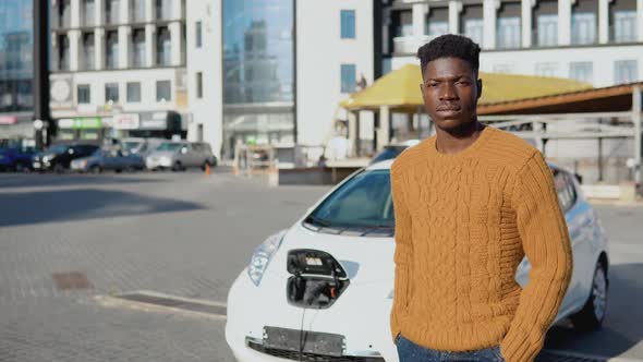 African American Driver is Standing Near a White Electric Car While Charging the Battery alt