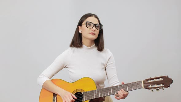 Woman In Beige Outfit Tuning Her Acoustic Nylon Strings Guitar On A Grey Background