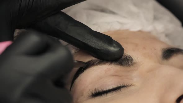 Close-up, the Hands of the Cosmetologist in Black Rubber Applying Permanent Make Up on Eyebrows- alt