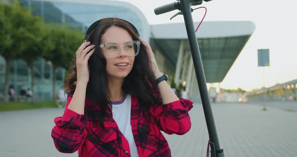 Woman in Stylish Clothes in Glasses Putting on Headphones while Sitting on E-scooter alt