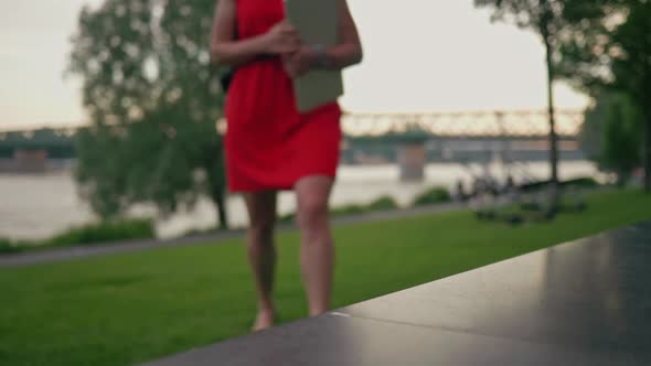 Beautiful woman in red dress with a laptop walking towards the bench in the park and sits on it. Cli alt