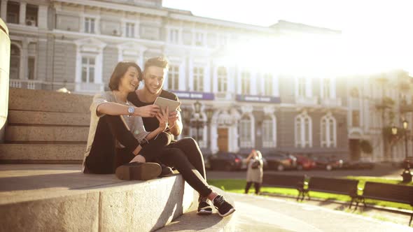 Young Beautiful Couple Smiling Speaking Looking at Tablet Sitting in City Park alt