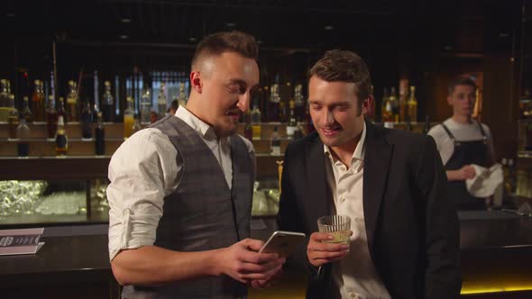 Two Men Stand at the Bar and Watch Photos on the Phone alt