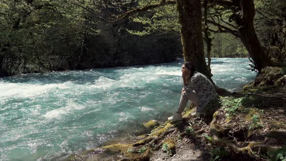 Young Woman Sits on the Shore Near the Swift River Stream She Looks at the Water alt