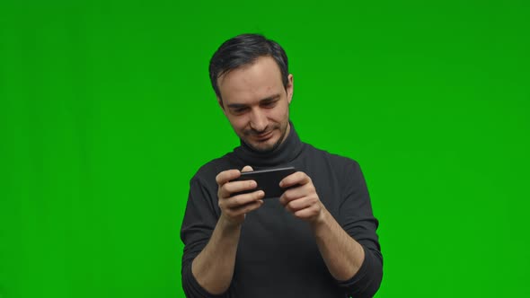 Excited Man in Sweater Playing Game on Mobile Phone Against Green Chroma Key Background alt