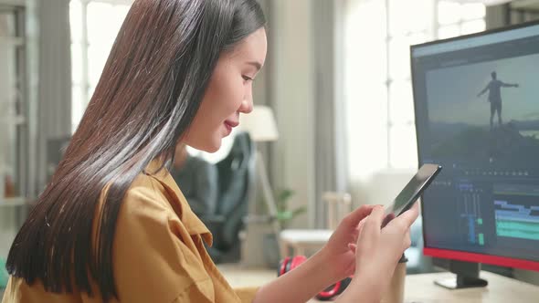 Asian Female Video Editor Works And Using Mobile Phone With Her Personal Computer In Office alt