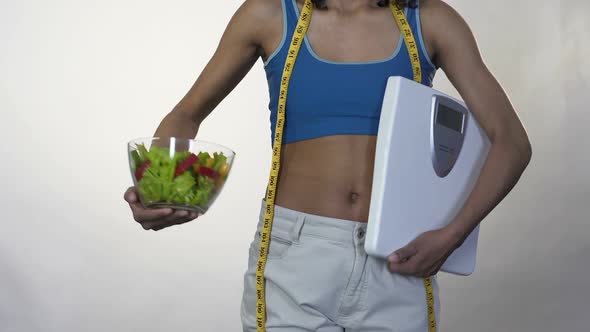 Woman in Loose Pants Standing with Scales, Holding out Bowl of Salad, Dieting alt