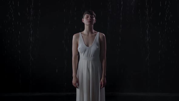 Lonely Sad Girl with Short Hair in a White Dress Stands in the Rain on a Black Background