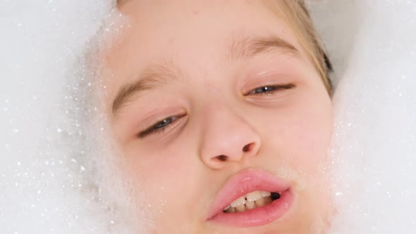 Portrait of Cheerful Teen Girl Lying in Bath with Thick Soap Foam and Looking in Camera