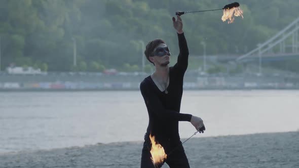 Young Slim Man in Black Clothes and Mask Performing a Show with Flame Standing on Riverbank alt