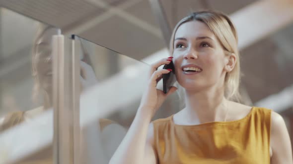 Young Blonde Businesswoman Leaning on Glass Wall and Chatting on Phone alt