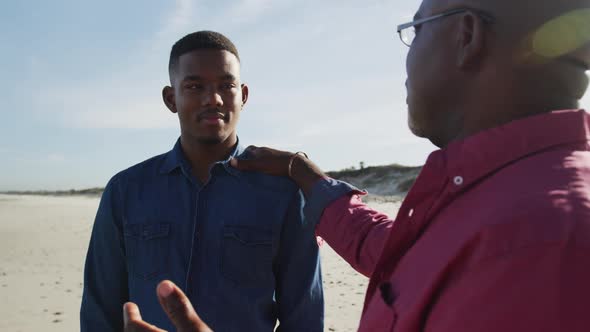 African american father standing on beach with teenage son, putting hand on his shoulder and talking alt