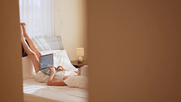 Smiling Woman Lying on White Bed While Lifting Legs Up and Using App on Laptop alt