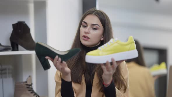 Thoughtful Millennial Woman Holding Casual Yellow Sneakers and Elegant Green Highheels Thinking alt