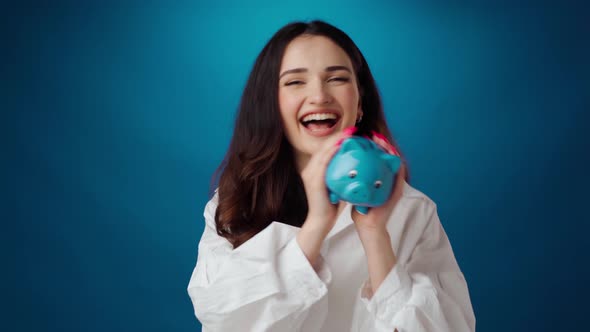 Pleased Young Woman Holding Piggy Bank Full of Money Against Blue Background