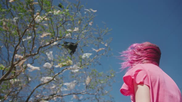 Back View of Hipster Woman with Pink Hairs at Green Tree Covered By Plastic Bags alt