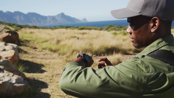 African american man hiking in countryside by the coast using smartwatch alt