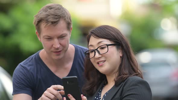 Mature Asian Businesswoman and Young Scandinavian Man Using Phone Together in the Streets Outdoors alt