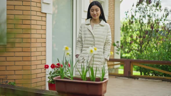 Happy Young Woman Walking to Blooming Flowers in Pot on Porch Smelling Fragrance alt