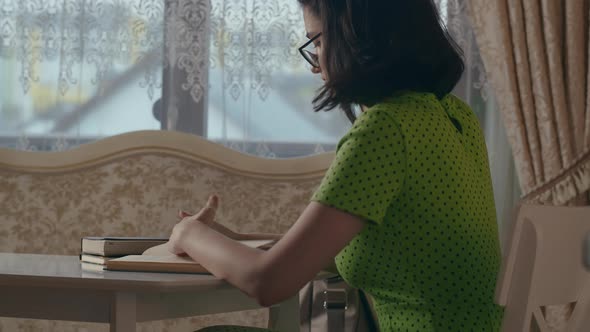 A Girl with Glasses and a Green Dress is Sitting at a Table and Reading a Book Side View alt