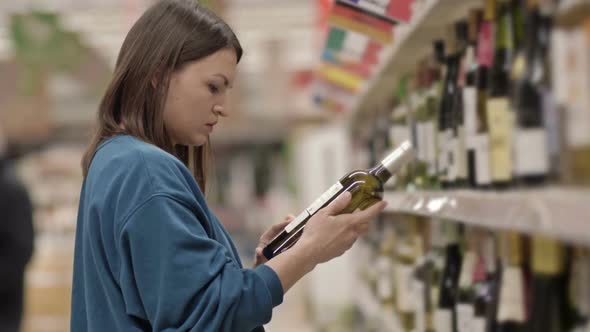 Young Woman Chooses Wine While Standing in Front of Shelves with Alcohol in a Supermarket or Liquor