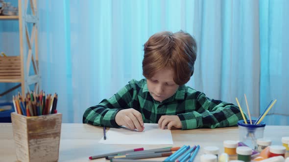 Little Schoolboy Sitting at Table in Room and Drawing with Pencil in Album alt