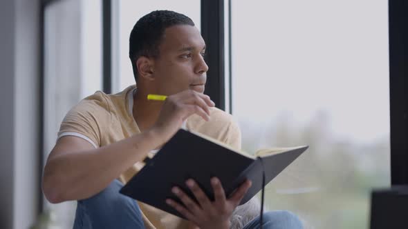 Portrait of Handsome African American Young Man Sitting on Windowsill Thinking and Writing in alt