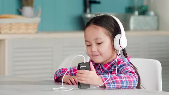 Little Asian Girl in Headphones Listening to Music on Smartphone