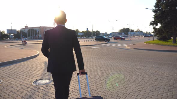 Rear View of Unrecognizable Young Businessman Walking to Airport Terminal and Pulling Suitcase on alt