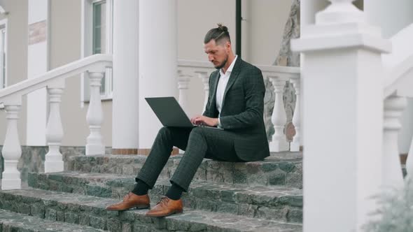 Handsome Bearded Businessman in Official Suit Is Working with Laptop Outdoor. Man Is Texting E-mail alt