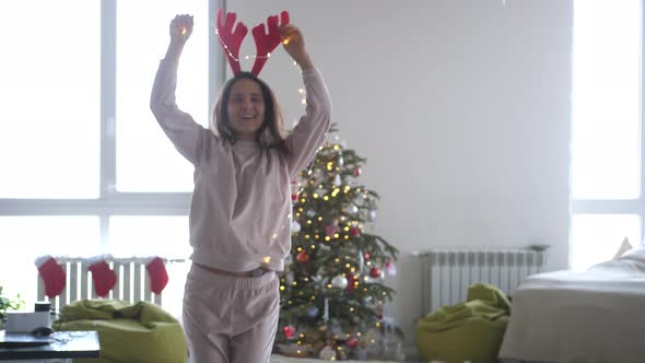 Young Beautiful Happy Woman Wearing Reindeer Antlers Headband and Garland Dancing Next to Christmas
