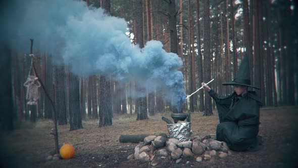 Terrible dark figure brews a potion in a cauldron in the forest. Scene for Halloween