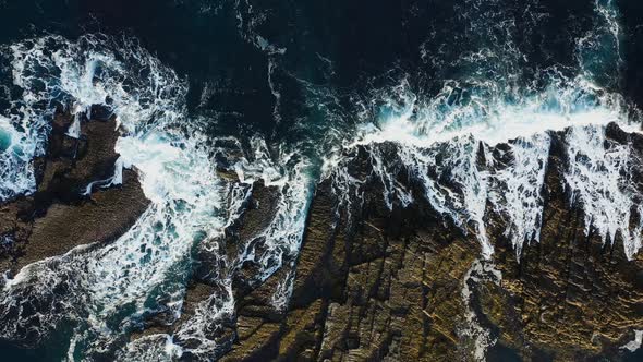 Sea waves with sea foam hit the rocky shore. Aerial cinematic top view.