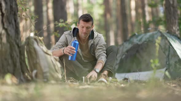 Front View Happy Male Tourist Pouring Drink From Thermos in Slow Motion Sitting at Campsite in alt