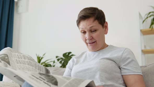 A Closeup View of a Beautiful Mature Woman is Reading a Newspaper Sitting on Sofa at Home in the alt
