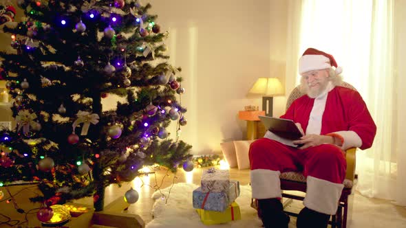 Laughing Positive Senior Bearded Santa Clause Gesturing Watching Online Movie on Tablet Sitting at alt