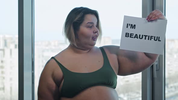 An Overweight Woman Holding a Nameplate with a Sign I'M BEAUTIFUL and Eating a Burger alt