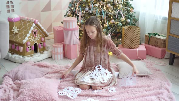 Adorable Little Girl Sitting Near the Tree and Making Paper Snowflakes