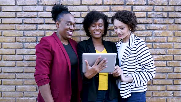 Three women make use of technology while standing outdoors, one of them points to the tablet alt