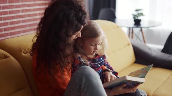 Little Girl Reading a Book with Her Mother While Sitting on the Sofa at Home alt