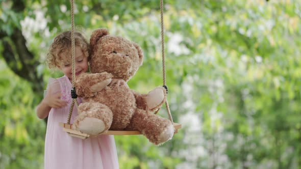 Spring and happy summer time. Joyful smiling little girl playing with teddy bear swinging on the swi