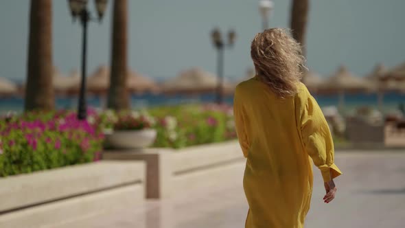 Blonde Woman in Yellow Dress is Walking to Sea Over Territory of Luxury Hotel alt