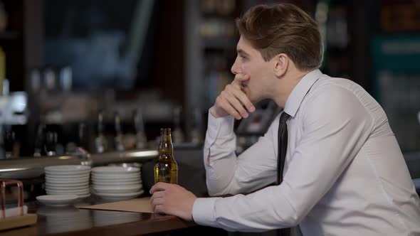 Side View Portrait of Frustrated Lonely Caucasian Man Drinking Beer From Bottle in Bar Indoors alt