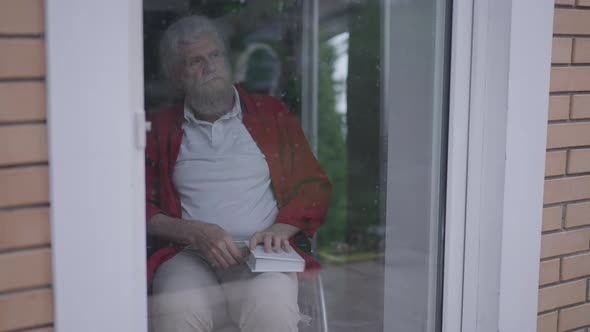 Depressed Thoughtful Senior Handicapped Man Closing Book Looking Out Window on Glass Door alt