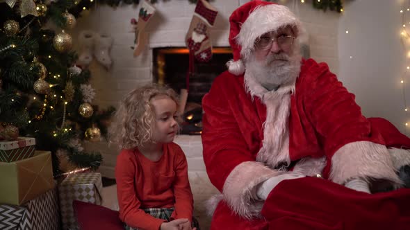 Funny Santa Claus Is Sitting on the Floor with a Little Cute Boy Sitting Next To Him, Opening a Gift