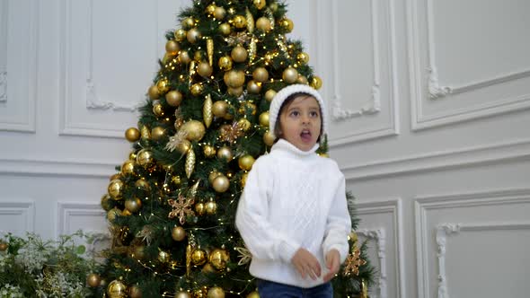 Boy Child in a White Sweater and Hat Stands at the Christmas Tree