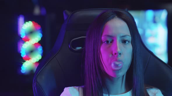 Girl Gamer Sitting in a Chair in Gaming Club and Blowing a Bubble Gum alt