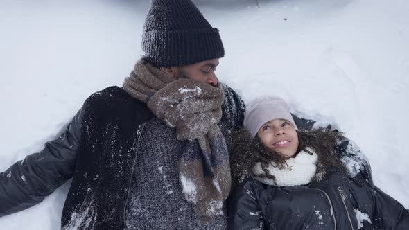 Carefree Laughing African American Man and Teen Girl Lying in White Snow Outdoors alt