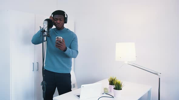 Man Wearing Headphones Singing a Song Into a Microphone in a Studio  Holding Phone alt